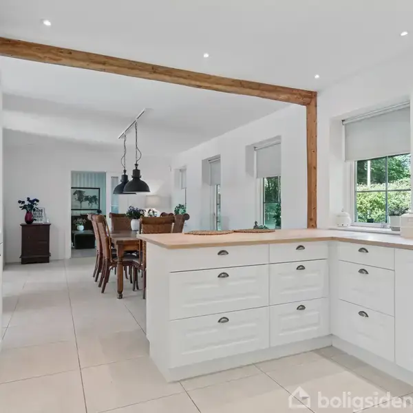 En lys køkken-alrum kombineret med spiseplads, domineret af hvide skabe og skuffer. Træbjælker opdeler rummet, med spisebord og stole i baggrunden. Vægtekst: "LOVE makes a house a home."