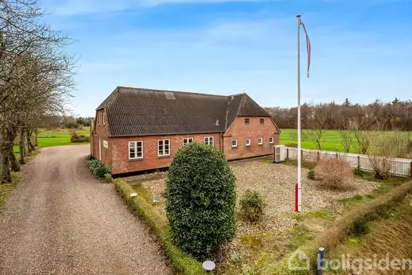 Hus i røde mursten står stille ved en grusvej, omgivet af en have med flagstang. I baggrunden ses en stor, grøn mark og træer under en klar, blå himmel.