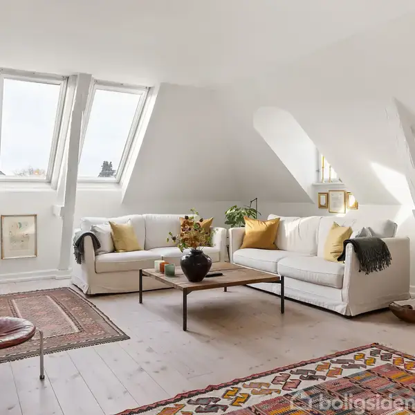 En lys stue med to hvide sofaer og et sofabord i midten. Store skråvinduer lader lys ind. Rummet er dekoreret med tæpper og planter, og kunst hænger på væggen.