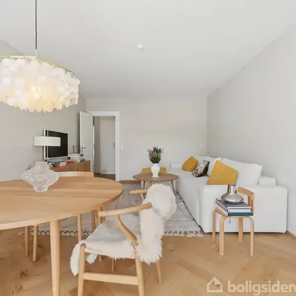 Lampe hænger over spisebord med fire stole i moderne, lyst indrettet stue med sofa, sofabord, fjernsyn og gulvlampe. Kaktus og hylder dekorerer rummet, der har træparketgulv.