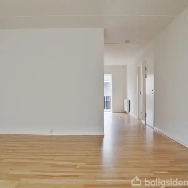Stue med trægulv og hvide vægge, set mod en åben korridor. Døre på højre side fører til andre rum, og en balkon kan skimtes i baggrunden.
