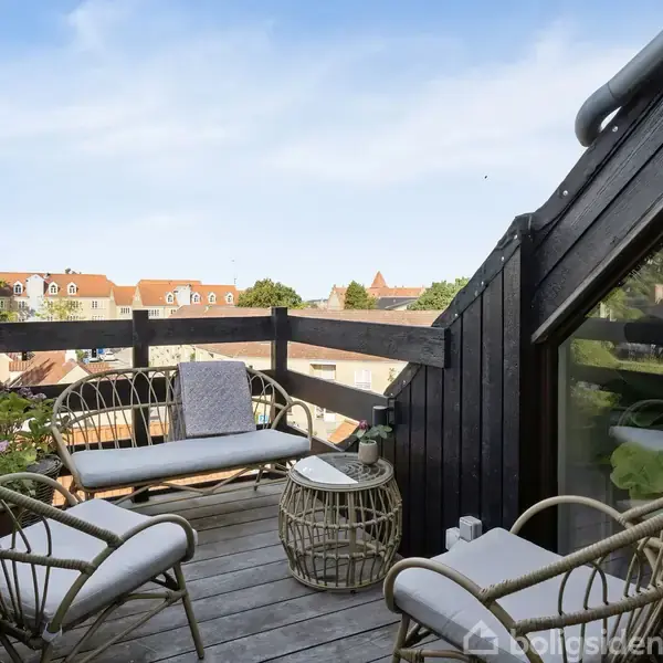 Terrasse med fletmøbler og bord, omkranset af sort rækværk. Udsigt til hustage og grønt. Klar blå himmel i baggrunden skaber en hyggelig og afslappet stemning.