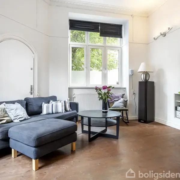 En blå hjørnesofa står i en lys stue med trægulv. Foran sofaen er et rundt sofabord med blomster. Der er et stort vindue, væglampe og fladskærms-TV på væggen.