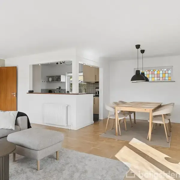 En lys stue med en grå sofa og sofabord står i venstre side. Til højre er der et spisebord med fire stole under en hængelampe. En åben køkkenområde ses bagved.