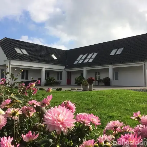 Et stort, hvidt hus med sorte tage står roligt i en grøn have med lyserøde blomster i forgrunden under en delvis skyet himmel.