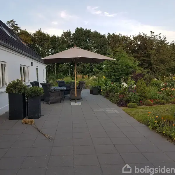 Haveområde med parasol og siddepladser på en flisebelagt terrasse. Til venstre ses et hvidt hus, og til højre er blomsterbede og buske. Skyfri himmel i baggrunden.