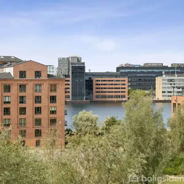 Røde murstensbygninger står statisk ved en flod med moderne højhuse i baggrunden, omgivet af frodige træer og buske under en klar blå himmel.