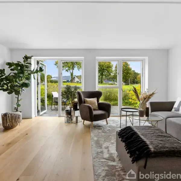 Stue med grå sofa, lænestol og sofabord, åbner mod terrasse gennem store vinduer. Lys træparketgulv, dekorative planter og udsigt til grønt område udenfor skaber en lys og indbydende atmosfære.