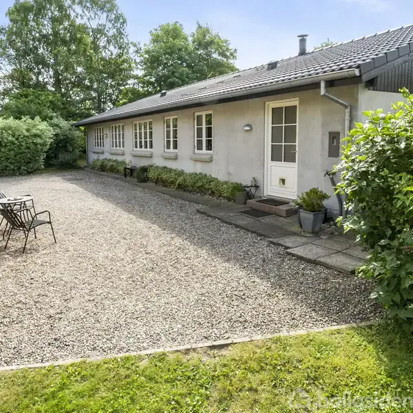 En enkelt etagers hus med hvid facade står langs en grusbelagt gårdsplads. To stole er placeret på gårdspladsen. Huset er omgivet af grønne buske og træer.