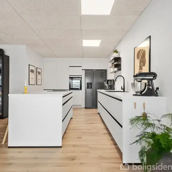 Et moderne køkken med hvide skabe og sort vandhane står centralt. En krukkeplante er på bordet i en stue med store vinduer. En hvid skammel står ved øen.