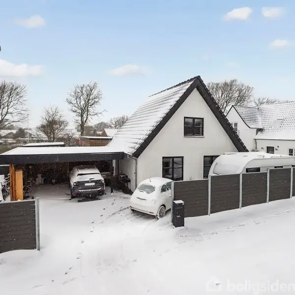 Et hvidt hus dækket af sne står stille. To biler er parkeret i indkørslen ved siden af en carport. Omgivet af et hegn og et vinterlandskab med træer.