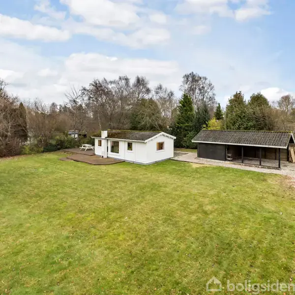 Hus i hvidt står stille ved siden af en carport, omkranset af en stor græsplæne og træer i en rolig, naturlig landsbykontekst.