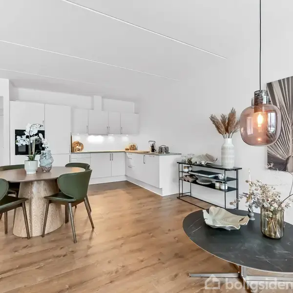 Køkken med hvide skabe og indbygget elektronik. Rundt spisebord med fire stole. Dekorative elementer inkluderer blomster og moderne kunst. Trægulv og store pendellamper til belysning.