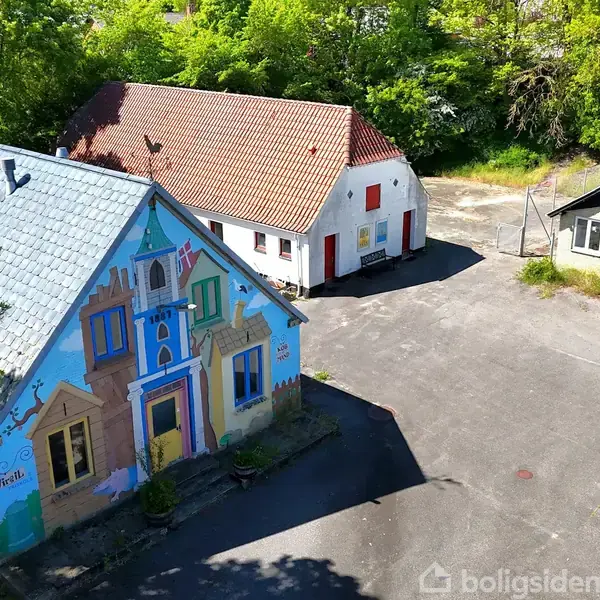 Et farverigt, dekoreret hus med en murmaleri af et fantasifuldt landskab. Det står på en asfalteret gårdsplads omgivet af flere mindre bygninger. Grønne træer omkranser området.