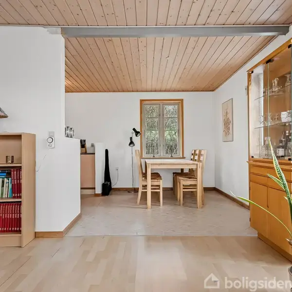 Bogreol med bøger står stille i stuehjørne. Et spisebord med stole er i baggrunden ved vinduet, planter til højre. Trægulv og loft skaber en varm atmosfære.