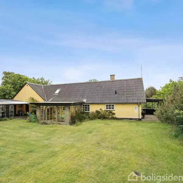 Et gult hus med mørkt tag står midt i en frodig have. Ved siden af ses en overdækning, og baggrunden består af grønne træer under en blå himmel.