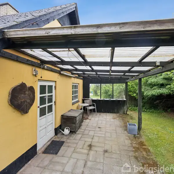 En sideliggende gul bygning med hvid dør og tilhørende overbygget terrasse. Terrassegulvet er flisebelagt, og omgivelserne har græs og træer. En dekorativ træudskæring hænger på væggen.