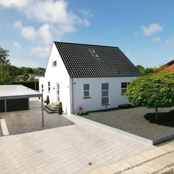 Et hvidt hus med sort tag står stille i en velholdt have med grusstier og et velplejet træ. Der er en garage til venstre og blå himmel.