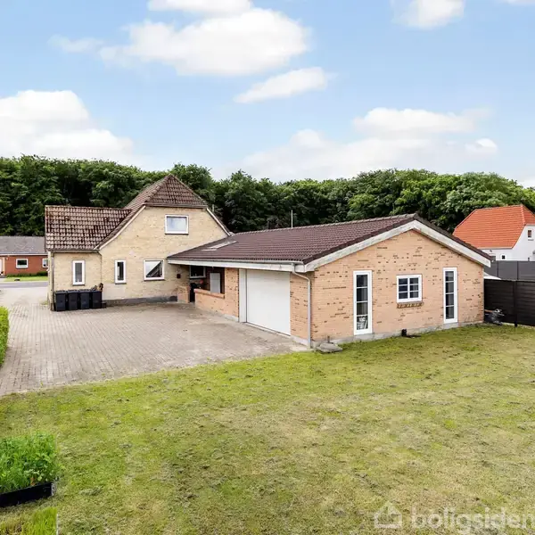 Rødt murstenshus med garage står stille på en flisebelagt gårdsplads, omgivet af en trimmet græsplæne og hække. Baggrunden er skovklædt og der ses flere lignende huse i nabolaget.