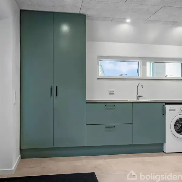 Et køkkenområde med grønne skabe til venstre står stille i lys belysning. En vask ved siden af vaskemaskine og tørretumbler. Et lille vindue over arbejdsbordet giver naturligt lys.