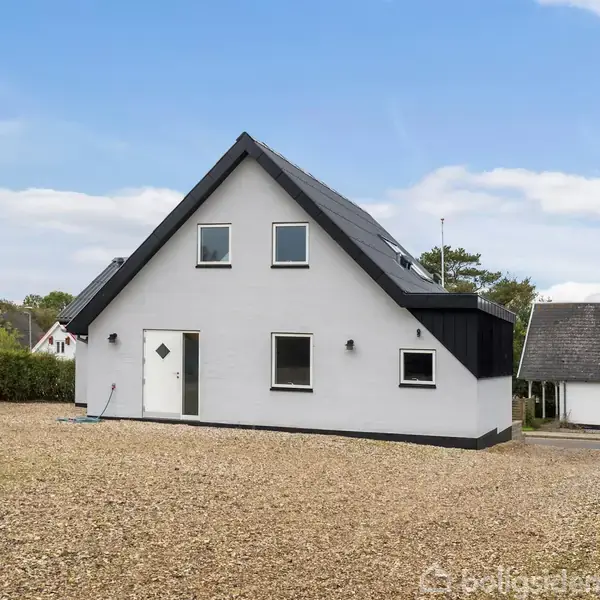 Et hvidt, trekantet hus står roligt med stejlt sort tag. Omgivet af en grusbelagt gård og grøn bevoksning under en blå himmel.