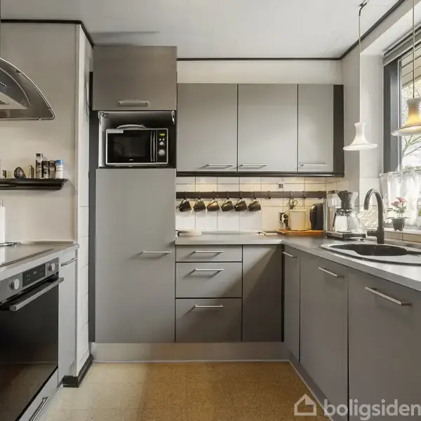 Køkken med grå skabe og hvide bordplader. En ovn og mikrobølgeovn indbygget i skabet. Bordet har køkkenredskaber, planter og en blender. Vindue ved vask med hvidt gardin.