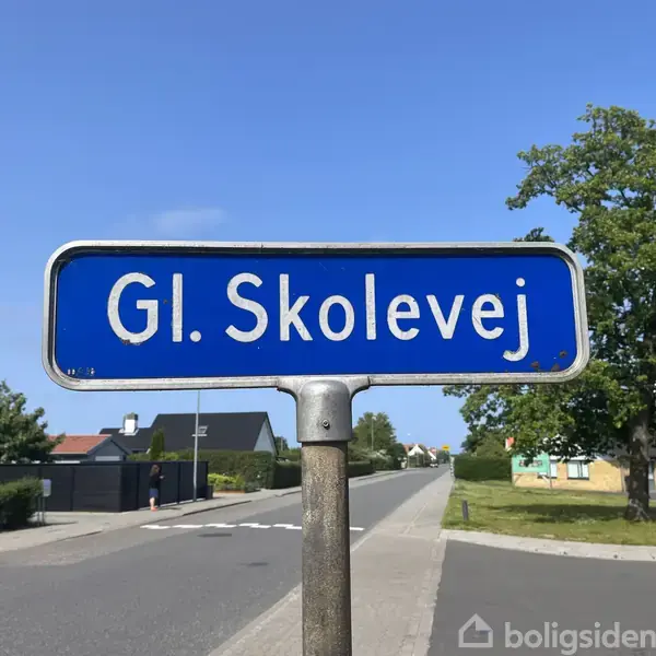 Et vejskilt viser teksten "Gl. Skolevej" på en blå baggrund. Det står i et beboelseskvarter med huse og en grøn træbevoksning i baggrunden.