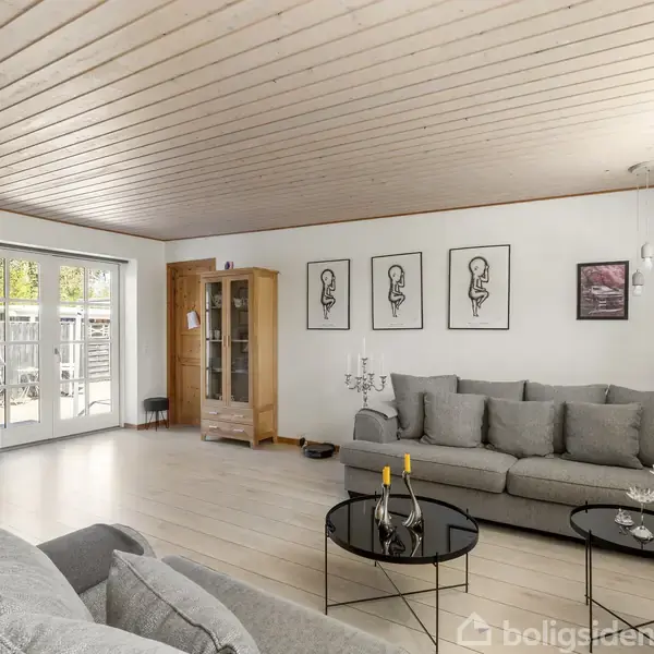 En stue med grå sofaer placeret ved et sofabord. En brændeovn står ved en hvid væg nær terrassedøre. Kunstværker hænger over sofaen, og et vitrineskab står i hjørnet.