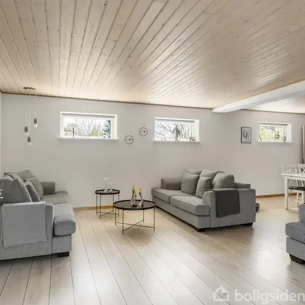 Stue med grå sofaer og glasbord viser moderne indretning. Rummets lyse træloft og trægulv skaber en åben atmosfære. Billeder pryder væggene, og spisebord ses i baggrunden.