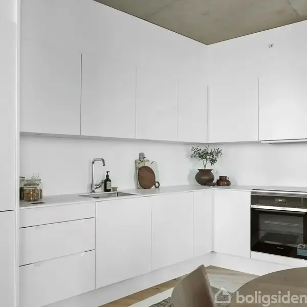 Et moderne køkken, hvor hvide skabe omgiver en indbygget ovn og vask. Dekorationer som planter og skærebrætter er placeret på bordpladen. Enkel og minimalistisk indretning.