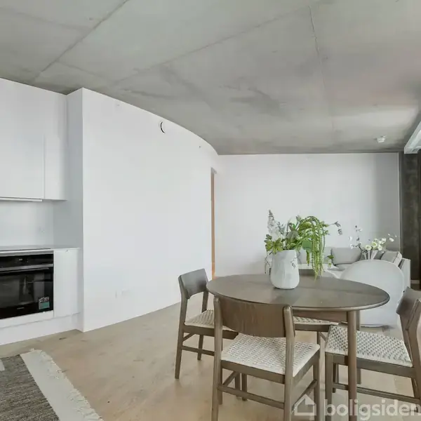Et rundt spisebord med fire stole står i et moderne, lyst køkken-alrum. Der er hvide køkkenskabe og sorte hvidevarer. Grønne planter står omkring rummet, som har lyse træplanker på gulvet.
