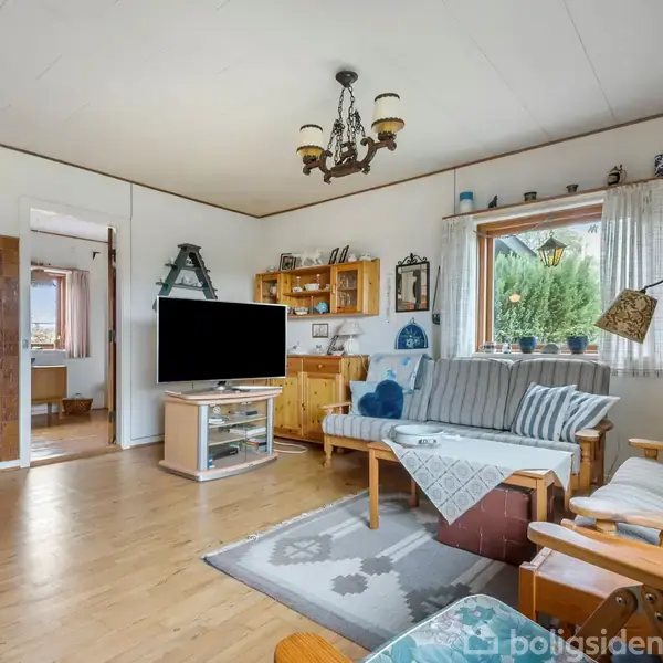 Brændeovn står tændt i en stue med træmøbler, sofaer og tv. Rummet er lyst med udsigt gennem store vinduer. Væggene har dekorationer og hylder med små genstande.