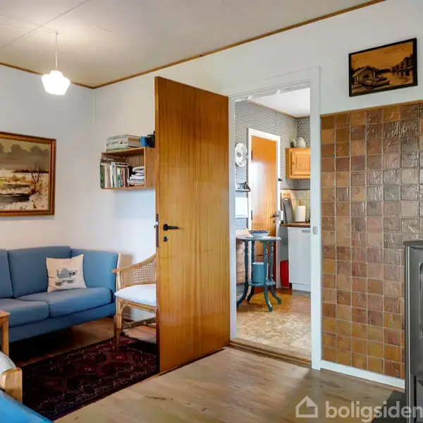 Brændeovn brænder i hjørne af stue ved siden af brun flisevæg. Blå sofa står foran træbord. Dør åbner til køkken med orange og træelementer i baggrunden.