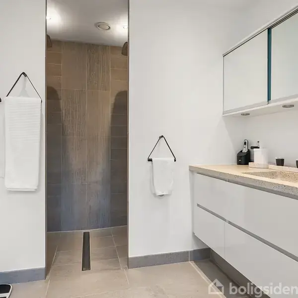 Et moderne badeværelse med en stor, hvid vaskeskab. Håndklæder hænger på væggen. En åben bruser er i baggrunden. Rummet er lyst og minimalistisk indrettet.