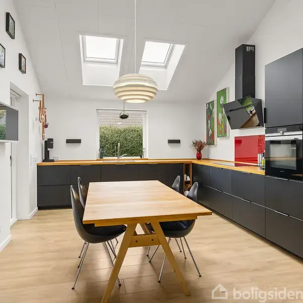 Et moderne køkken med sorte skabe og en træbordplade. En træspisebord med sorte stole står i midten. Ovenlys og vinduer giver naturligt lysindfald. Kunst hænger på væggene.