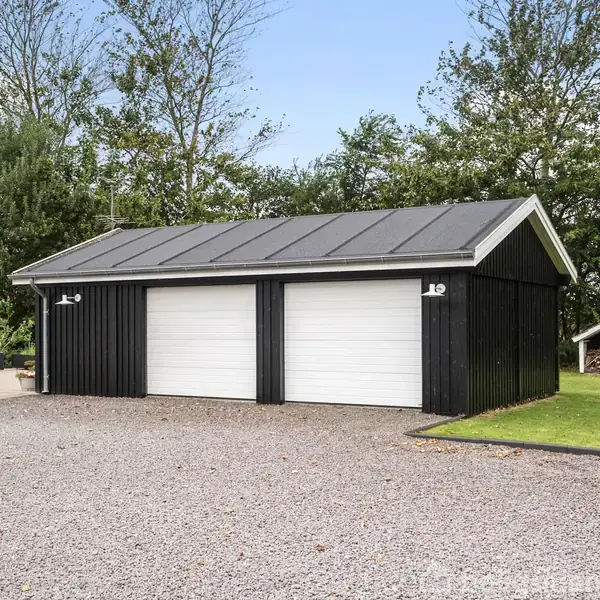 To-dørs garagebygning står på en grusbelagt indkørsel ved siden af en hvid bygning. Omkring garagebygningen er der grønne træer og buske, samt en lille mørk legehytte i baggrunden.