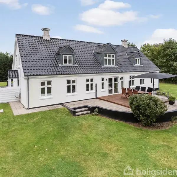 Hvidt hus med sort tegltag, stor terrasse med bord og parasol. Omkranset af grøn græsplæne og træer i en landlig have.