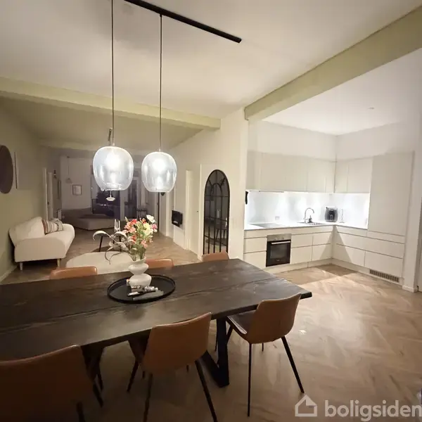Spisebord med stole står i et åbent køkken-alrum. Køkkenet er hvidt med indbygget ovn og vask. I stuen ses en sofa, sofabord og to store hængelamper over bordet.