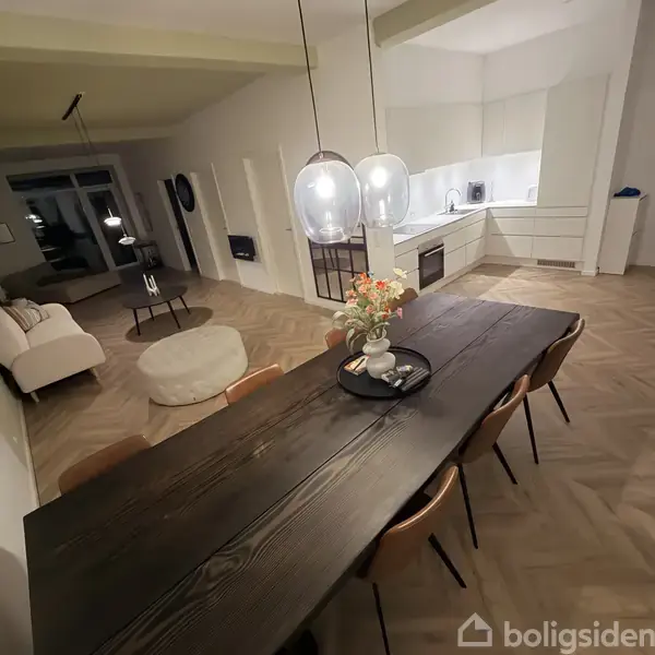 Langt spisebord i træ med seks stole og blomsterdekoration, beliggende i en åben moderne stue-køkken. Køkkenet har hvide skabe, og der er en sofagruppe til venstre.