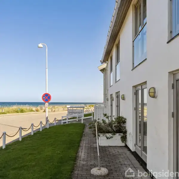 Bygning med store vinduer står stille langs en kystvej nær strand og hav, omkranset af græsplæne, stenbelægning og vejskilte med teksten "L22 35 m/herfra".