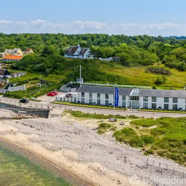 Bygning ligger langs en kystlinje, omgivet af en stenet strand og frodig, grøn natur. Flere små huse ses i baggrunden, hvor landskabet er bakket.