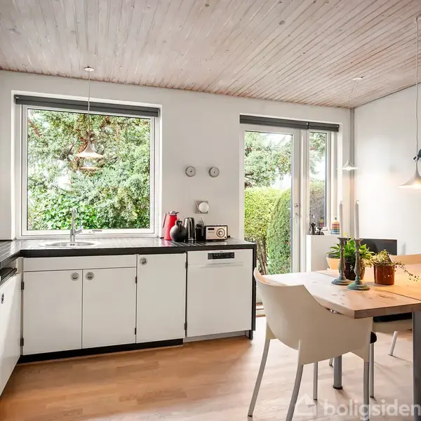 Køkken med hvide skabe og sort bordplade, ingen synlig aktivitet. Det er oplyst af naturligt lys fra store vinduer, og spisebord med fire stole ses i hjørnet.