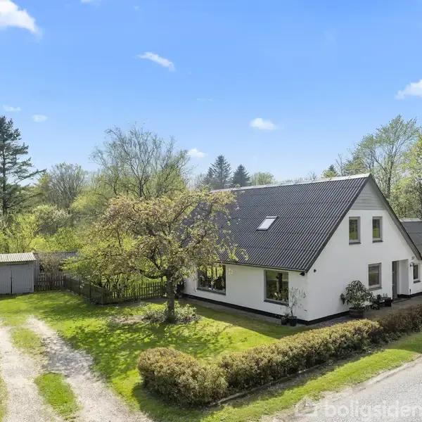 Hus med sort tag ligger ved en grusvej omgivet af grønne træer og buske. Ved siden af huset ses en garage. Klar himmel og sollys i baggrunden.