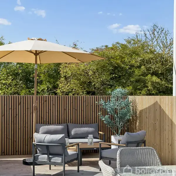 En terrasse med grå sofaer står under en parasol. Baggrunden domineres af et plankeværk og grønne buske, hvilket skaber et hyggeligt udemiljø.