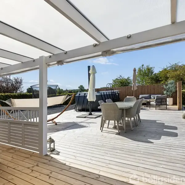 En stor træterrasse har en hængekøje og spisebord med stole under en hvid pergola. Havemøbler står i baggrunden omgivet af buske og hegn i en solrig have.