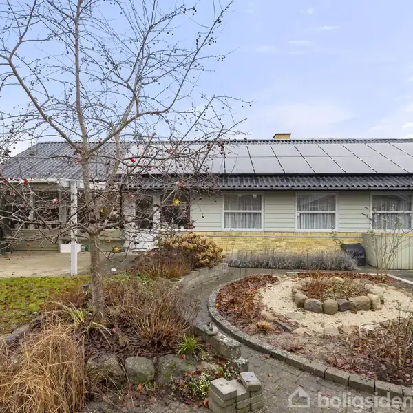 Et gult murstenshus med solpaneler på taget omkranses af en have med en sandkasse omgivet af en stenkant. Bar træstruktur i forgrunden. Himmel er klar og blå.