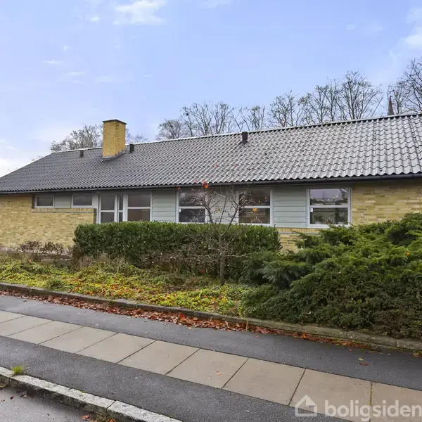 En gulstensvilla med mørkt tag står i et beboelseskvarter. Foran er der en have med buske. En bred fortov løber langs huset.