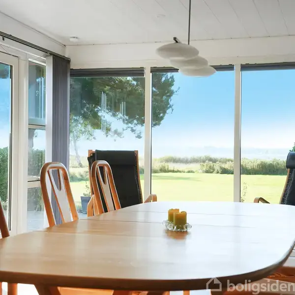 Bord med seks stole står i en lys stue. Store vinduer viser udsigt til en grøn have og blå himmel. Tre lamper hænger over bordet med tre gule stearinlys.