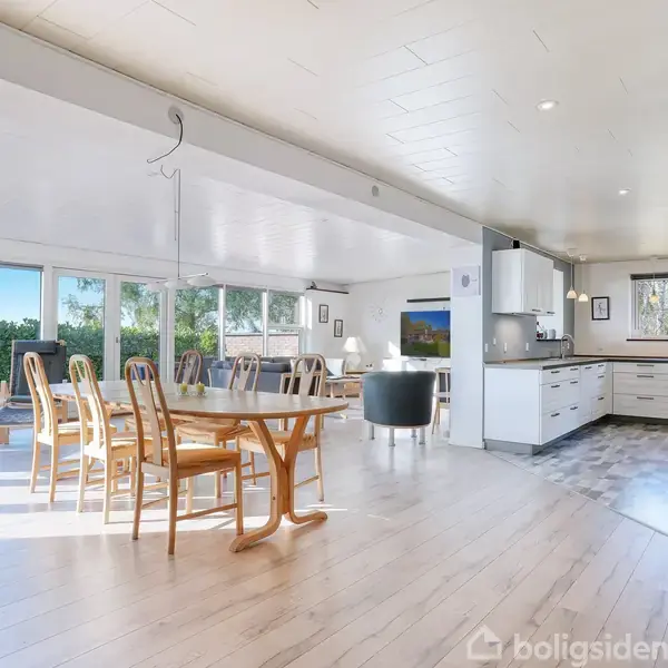 Spisestue med træbord og stole står i et lyst, moderne rum med store vinduer, der viser en grøn have. Køkkenet har hvide skabe, og stueområde ses i baggrunden.