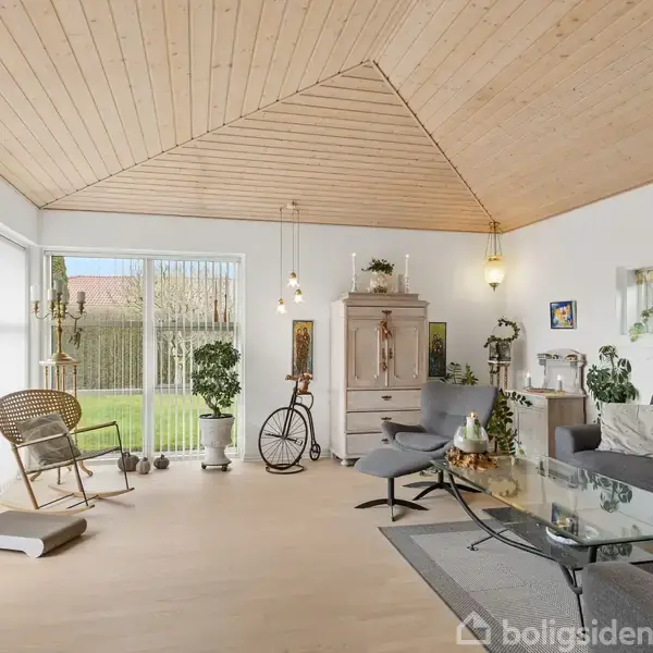 Stue med glasbord og grå sofa, dekoreret med planter og lyskilder. Store vinduer giver lys og udsigt til have. Trævægge og loft skaber en lys og hyggelig atmosfære.